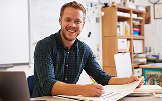 Smiling male teacher grading papers at his desk in a classroom