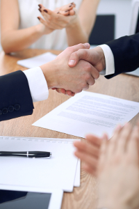 Close-up of a handshake over an office desk with documents indicating a business agreement or partnership deal, with additional people in the background.