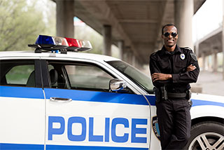 Confident police officer in uniform wearing sunglasses, standing with arms crossed next to a patrol car.