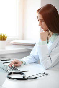 Young woman resting her chin on one hand while using an open laptop on a white desk with a stethoscope on it in a sunlit room
