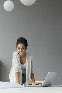 A smiling young woman with one hand leaning on a sheet of paper and the other hand resting on a laptop keyboard