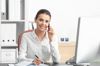 A smiling woman sitting at an office desk writing in a notebook and holding a phone to her ear