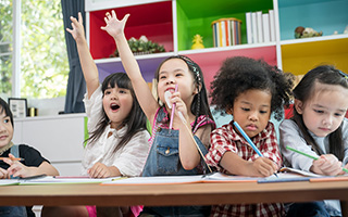 Two children with raised hands sitting at a table next to two other children writing in notebooks