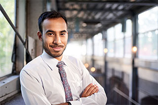 Smiling businessman in a white shirt and tie, standing with arms crossed in a modern office setting.