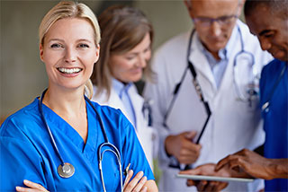 Smiling female nurse in blue scrubs with a stethoscope, standing confidently in front of a team of medical professionals.