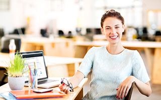 A smiling female office worker holding a pair of glasses and sitting at a desk with books, pens, a plant, and an open laptop
