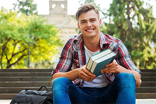 Smiling young male student sitting outdoors on campus steps, holding books.